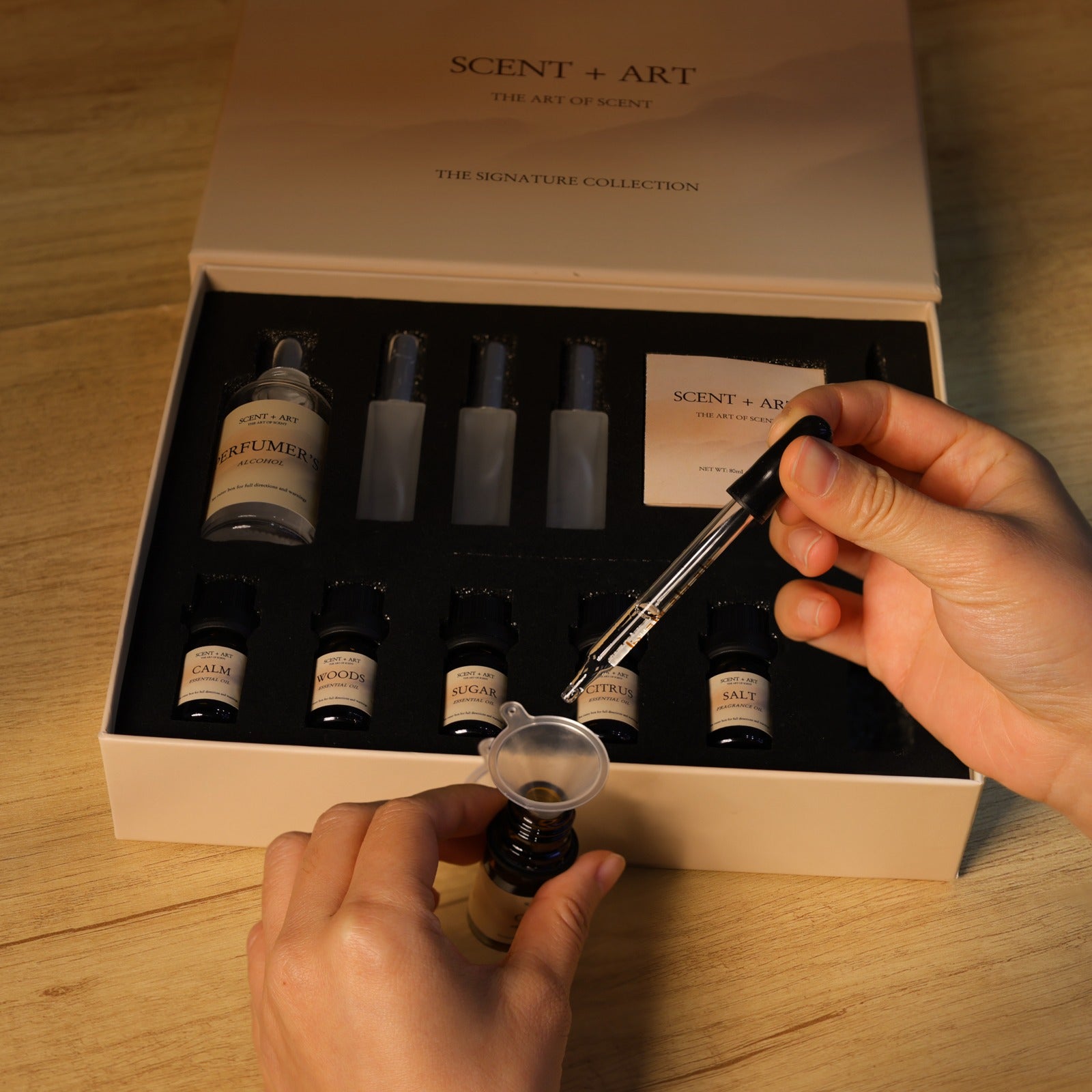 Hands using precision dropper to mix fragrance oils from SCENT PLUS ART DIY perfume kit, demonstrating the perfume-making process with open Signature Collection box showing organized components